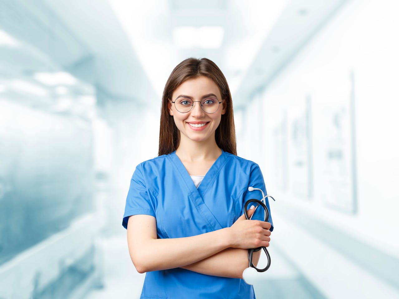 services-03 Young nurse in blue scrubs smiling and holding a stethoscope in a hospital corridor.