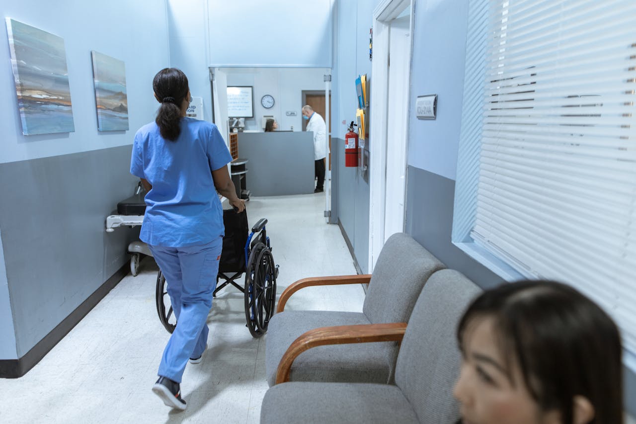 services-02 A nurse in scrubs pushes a wheelchair through a well-lit hospital corridor, attending to patient care.