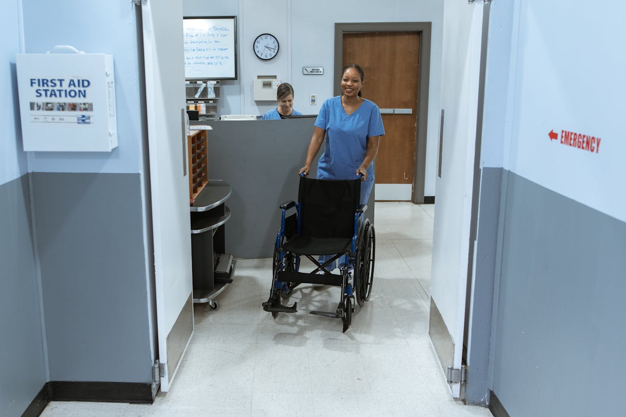 Home A nurse in scrubs pushes an empty wheelchair through a hospital hallway.